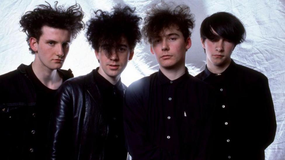 Jesus and Mary Chain. Four men wearing all black. Three of them have backcombed dark hair while the fourth has his dark hair swept over his face, obscuring his eyes 