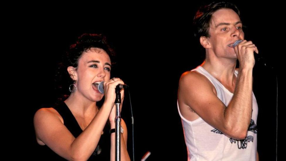 Lorraine McIntosh and Ricky Ross singing on stage during the eighties. They are both wearing vest tops.