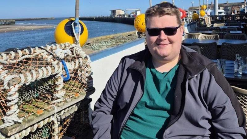 Anthony Maines sitting in a seaside restaurant. He is wearing sunglasses, a green t-shirt, and a jacket. Behind him, you can see the docks, the sea, and the blue sky.