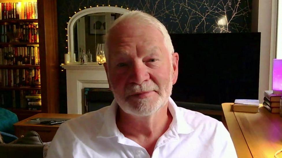 Tim Riesner sits in his living room with a desk next to him and a fireplace behind him.