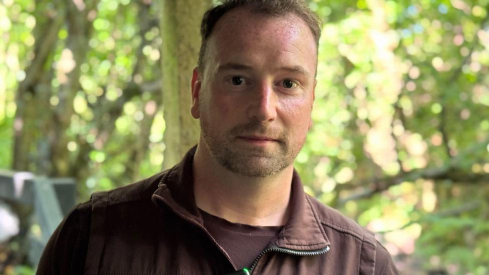 Peter Mitchell has short light brown hair and a beard. he is wearing a brown zip up gilet over a brown t-shirt and is standing in a wooded area.