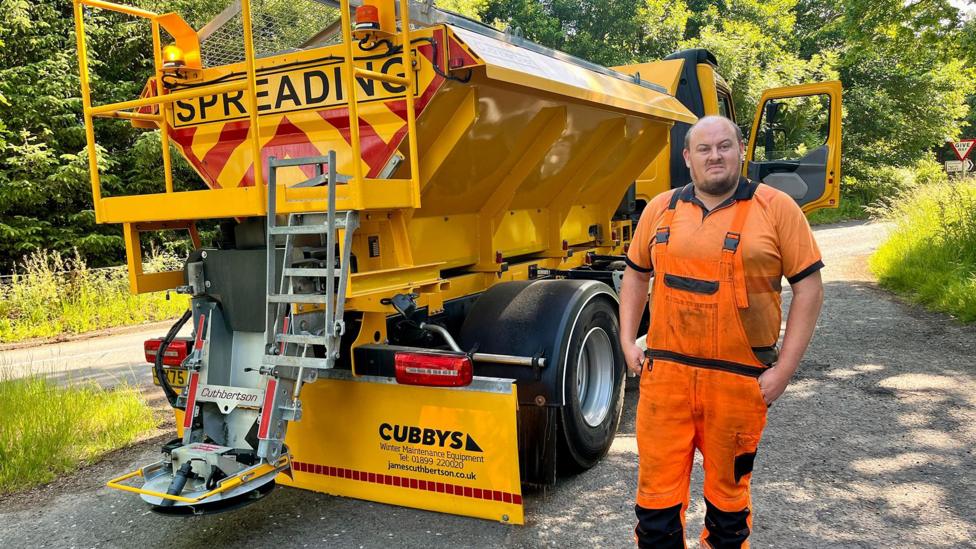 Road worker in orange high-visibility clothing standing beside a yellow gritting truck marked Spreading on a rural roadside with trees in the background.
