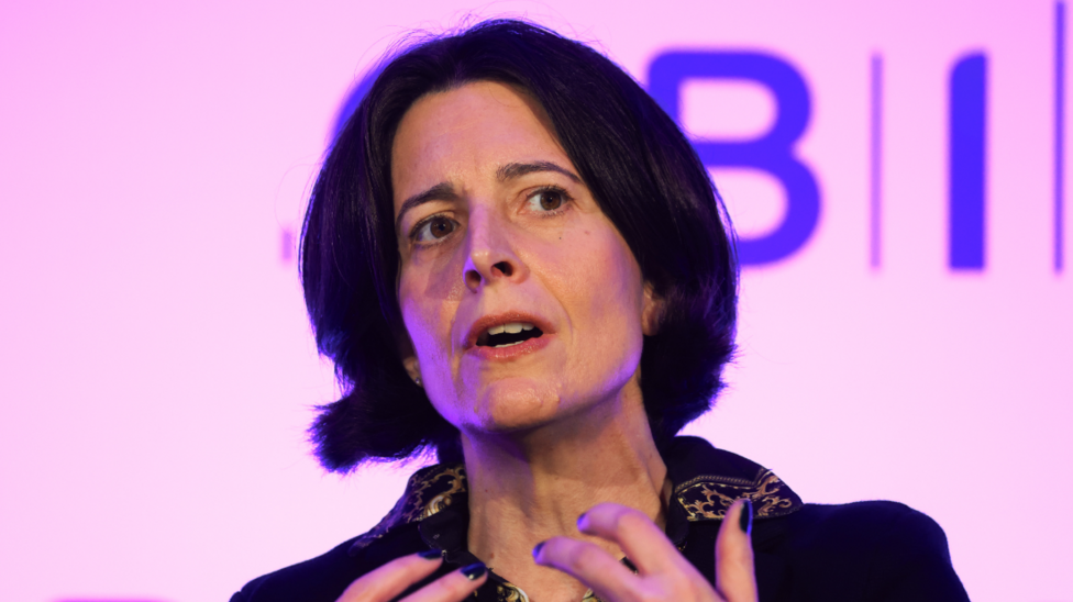 A woman with short black hair is speaking on a stage. She is sitting down and is wearing a black jumper. 