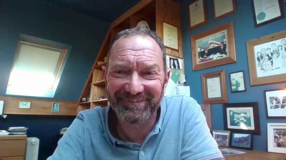 David Blevings is a middle aged man with short, greying brown hair and a beard. He is wearing a blue polo-shirt and is smiling at the camera. Behind him is an office with the walls painted blue and lots of pictures on on them. 