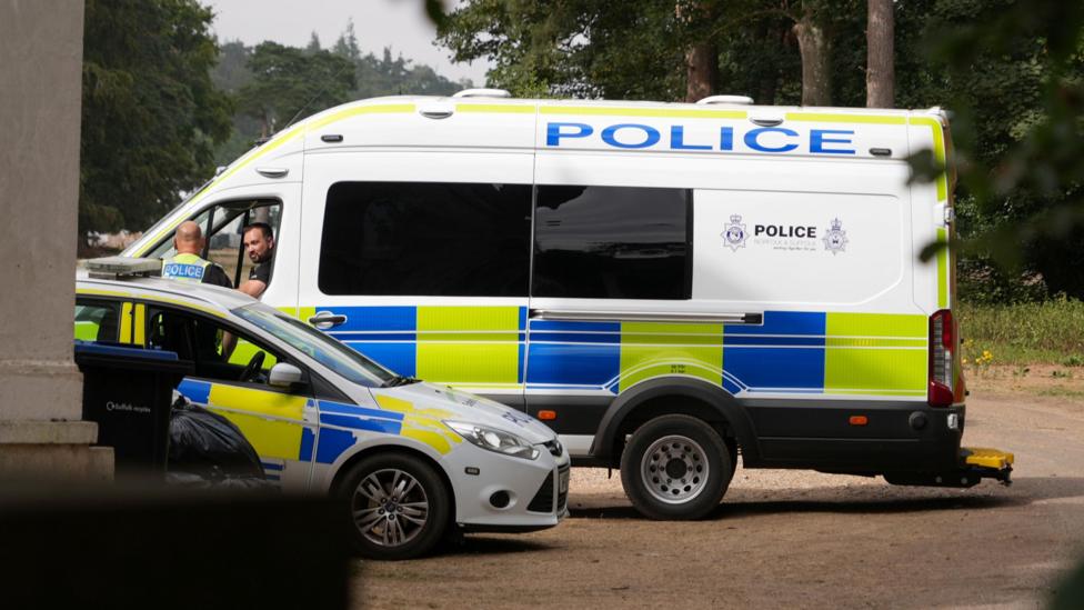 A police van and a police car parked next to each other. Two officers – one in the passenger seat of the van and one standing outside – are talking.