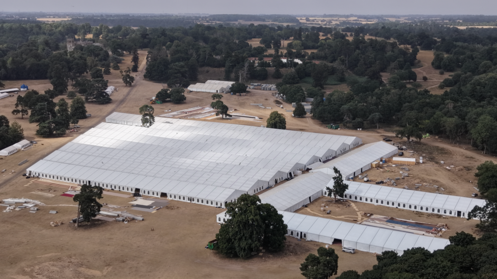 An aerial photograph of a massive complex of interconnected white marquees and tents set up in a large, open field surrounded by dense woodland.