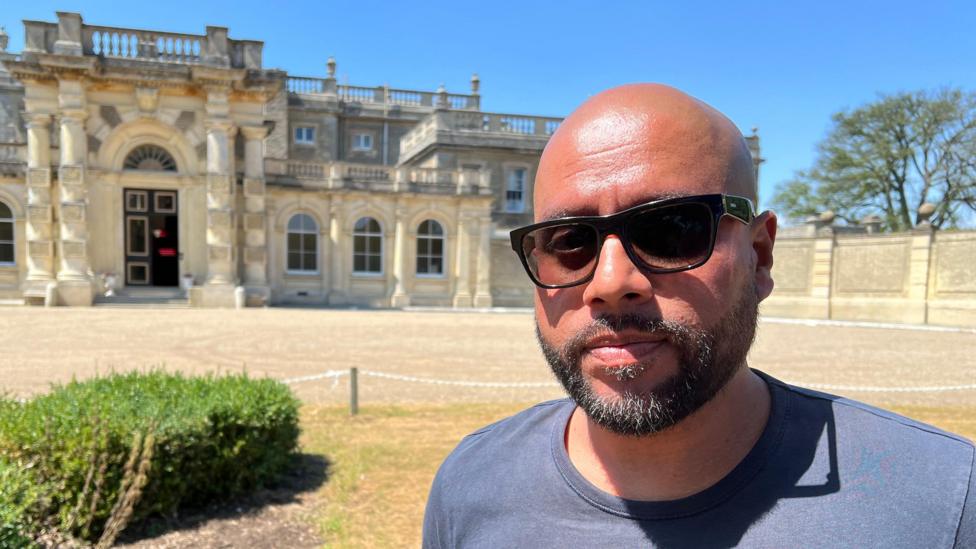 A man wearing sunglasses and a T-shirt while standing in front of a grand country house.