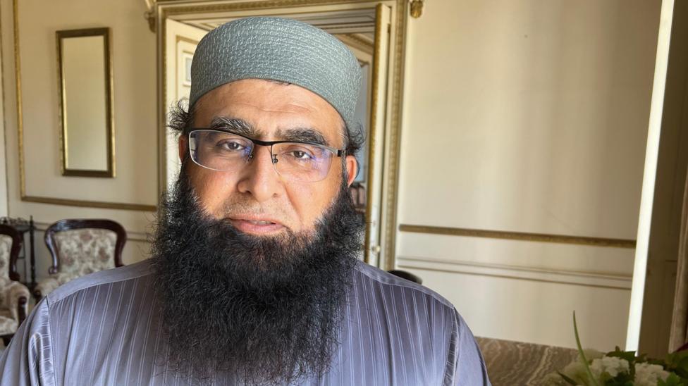 A man with glasses and a long black beard. He is wearing a hair covering and looking into the camera.