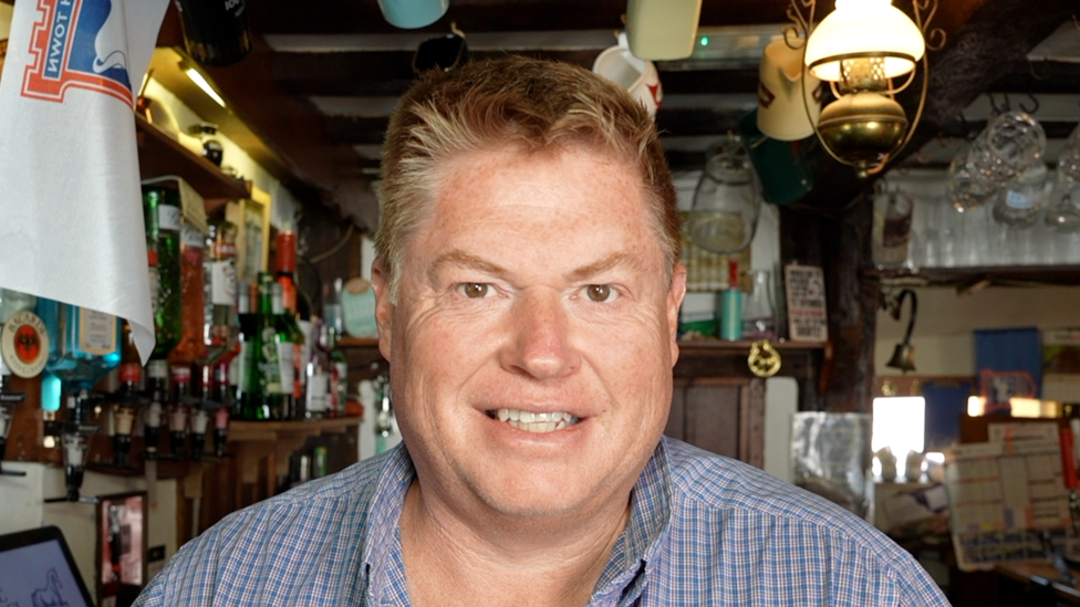 A man standing behind a bar. He is wearing a check shirt and smiling at the camera.
