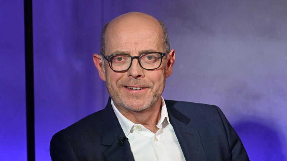 Nick Robinson is smiling at the camera. He is wearing a white shirt and a navy suit jacket. There is a purple background.