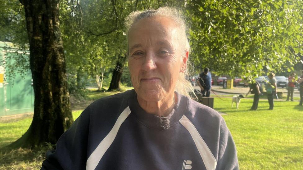 A woman standing in a park with a blue sweatshirt on. She has grey hair.