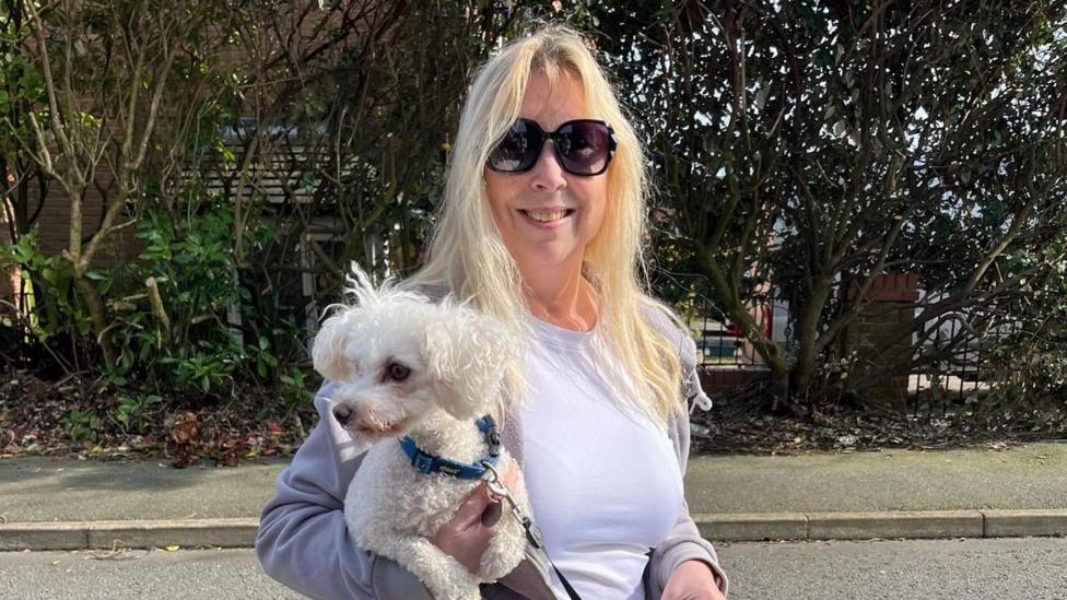 Sheena, a blonde lady wearing sunglasses, is holding a small fluffy white dog, Henry, and smiling.