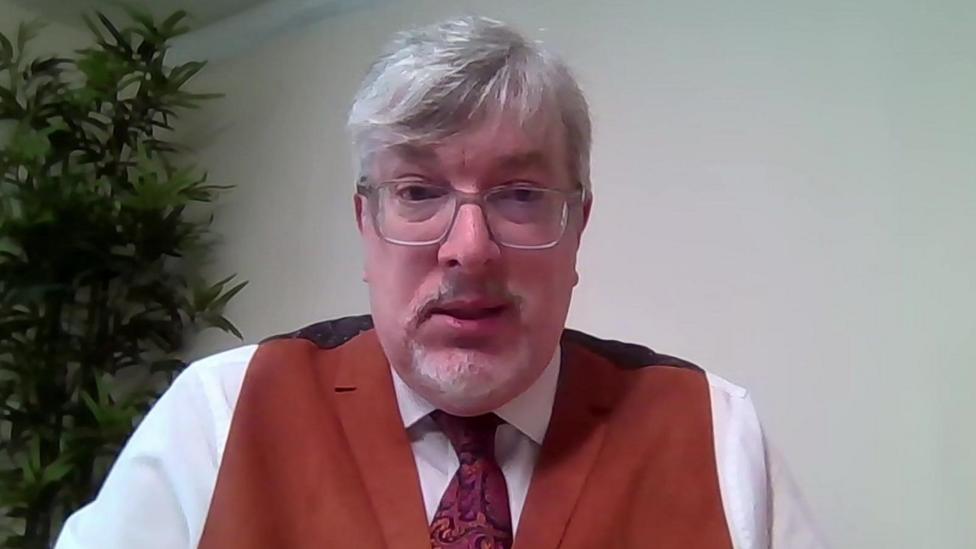 Man with grey hair and a grey beard wearing clear-framed glasses, a white shirt with a pink patterned tie and a red waistcoat.