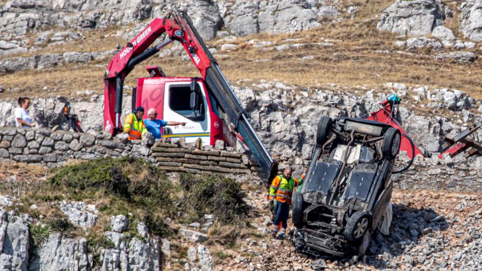 Upside down car pulled back to road above where a lorry with long arm and winch attempts to recover it