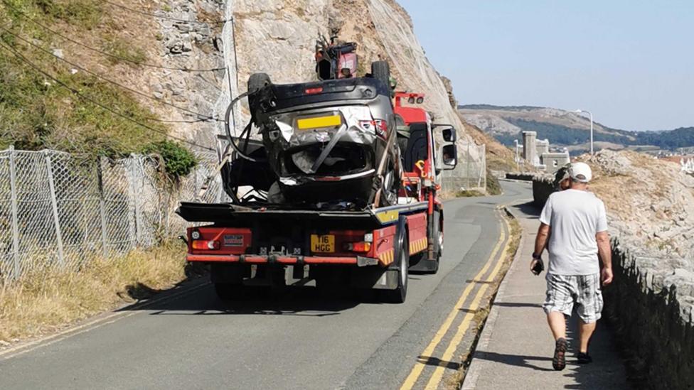 Upturned silver car fixed to back of red low loader and driven away as passersby look on