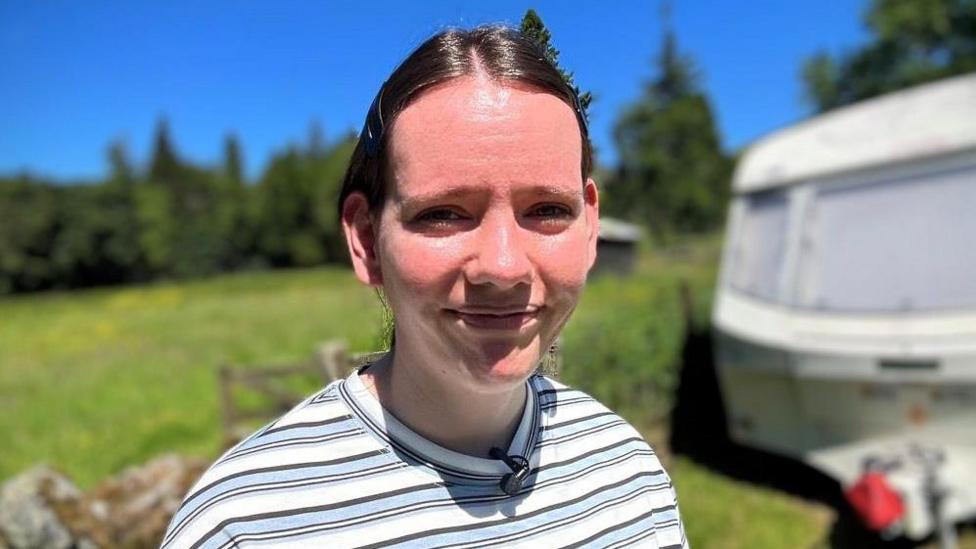 Samantha Donaldson has dark hair in a low ponytail. She is smiling at the camera into the sun with a caravan and grassy field blurred in the background.