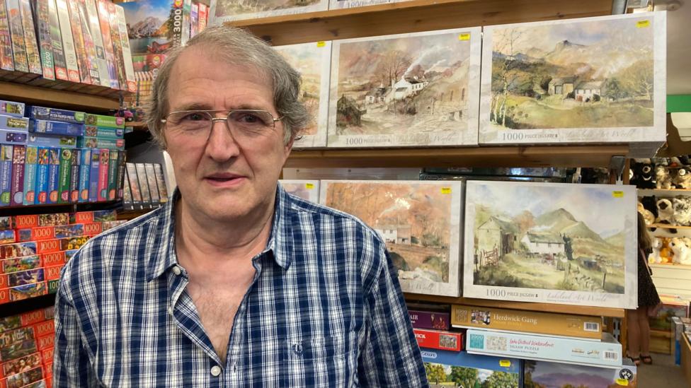 John Lowther standing on from of lots of jigsaw puzzles, many showing countryside paintings with mountains and trees in them. He has grey hair and is wearing a blue and white checked shirt.