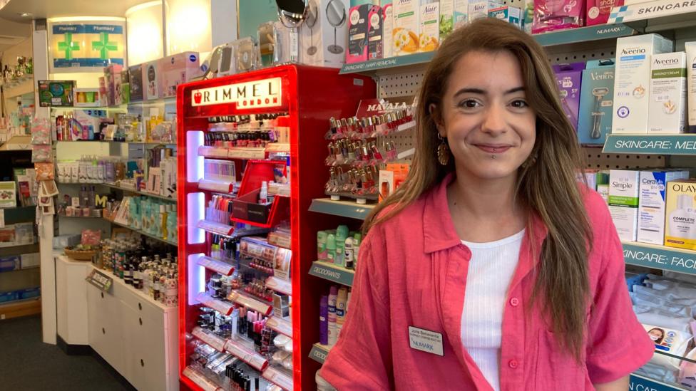 Jone Benevente has long brown hair. She is wearing large earrings and smiling. She is wearing a pink shirt and has a name badge on. In the background there are shelves with makeup and other pharmacy items.
