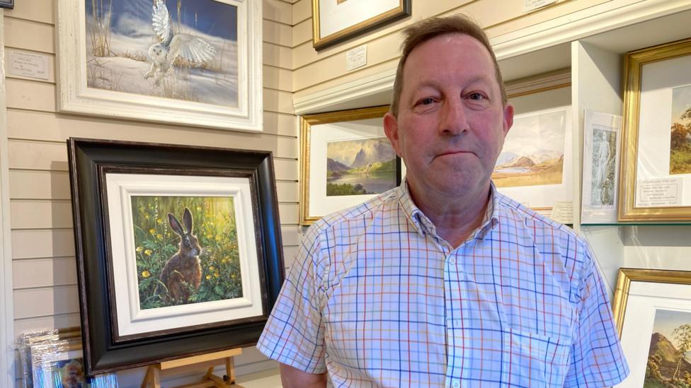 Tim Hardy has short brown hair. He is wearing a checked shirt. Behind him to the left there is a framed painting on an easel. It shows a rabbit or hare in a field with green flowers. There are other paintings in the background.