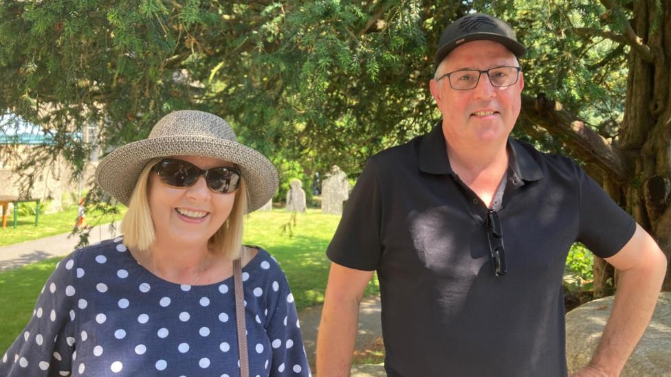 Glenda Grey is smiling and has blonde hair. She is wearing a hat, sunglasses and a blue and white polka-dot top. On the right, Phil Grey is wearing glasses, a black polo shirt and baseball cap. Behind them there is a graveyard. It is sunny.