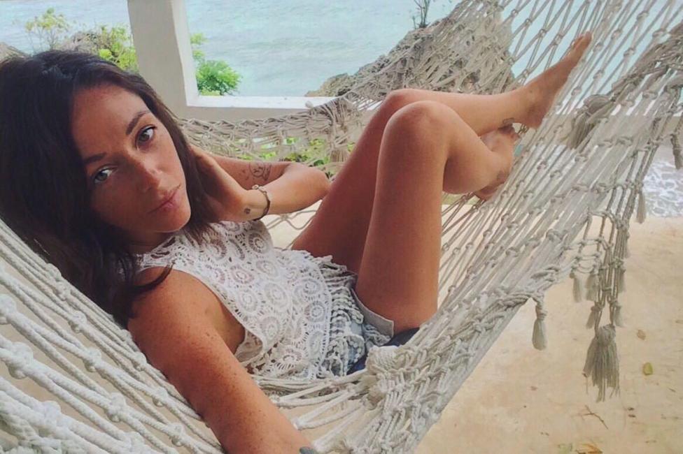 Kirsty McKie lies in a hammock and looks at the camera as she takes a selfie. The sea and a tree can be seen in the background and sand below her. She has brown hair that touches her shoulders.
