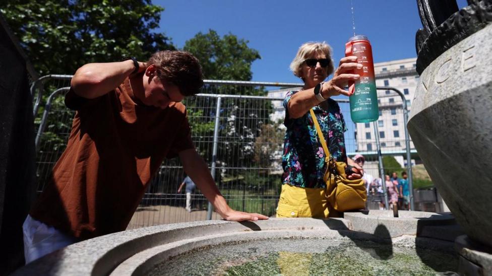 A man leaning over a fountain to pour water on his neck. A woman holding a water bottle under the fountain tap.