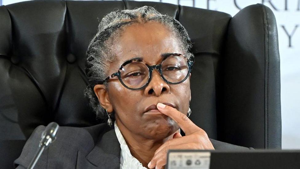 Sesi Baloyi, wearing black framed glasses and a grey suit jacket, holds a finger to her lips