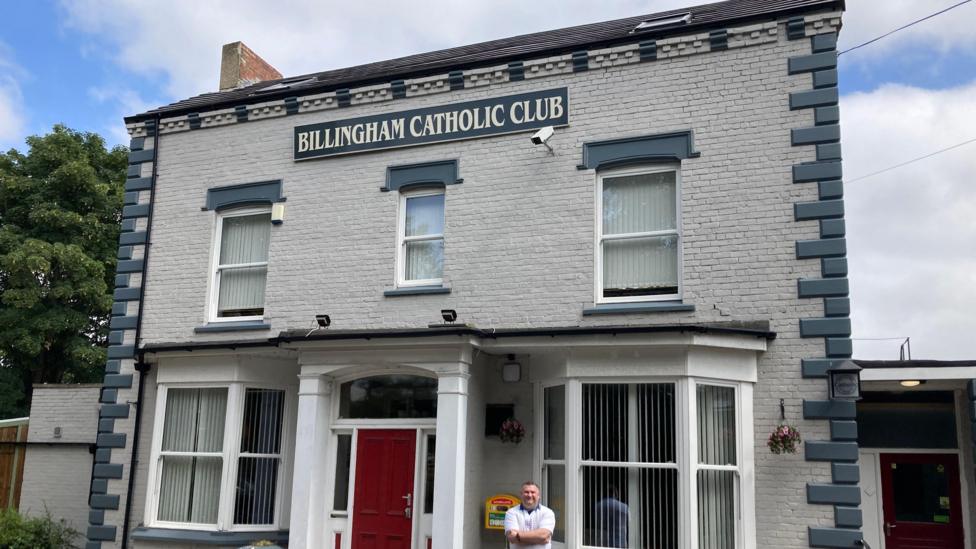 The exterior of the Billingham Catholic Club. The facade is mostly white with some blue accents. It has a bright red door in the middle and a brown door on the right hand side. The building is two storeys. Sean Rowbottom is standing, smiling at the foot of the building with his arms crossed.