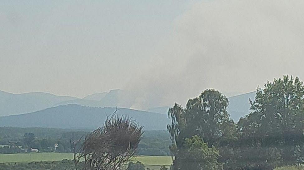 A countryside scene surrounded by green trees and smoke billowing from the hills
