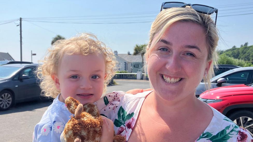Nicole O'Neill has her blonde hair tied back and is wearing sunglasses on top of her head. She is wearing a colourful floral dress and is holding her daughter, Holly, in her arms. Holly is holding onto a teddy and has short, curly blonde hair.