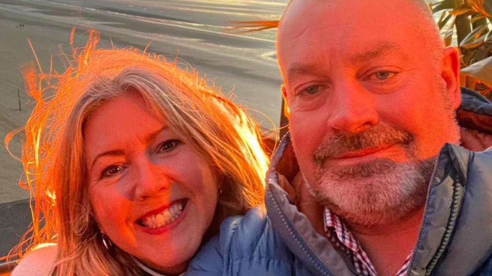 Joanne Brandon-Hodgkinson with David Griffiths. Joanne has shoulder length blonde hair. Griffiths has cropped grey hair and a beard, He is wearing a checked shirt and a puffer jacket. They are smiling and by the sea.