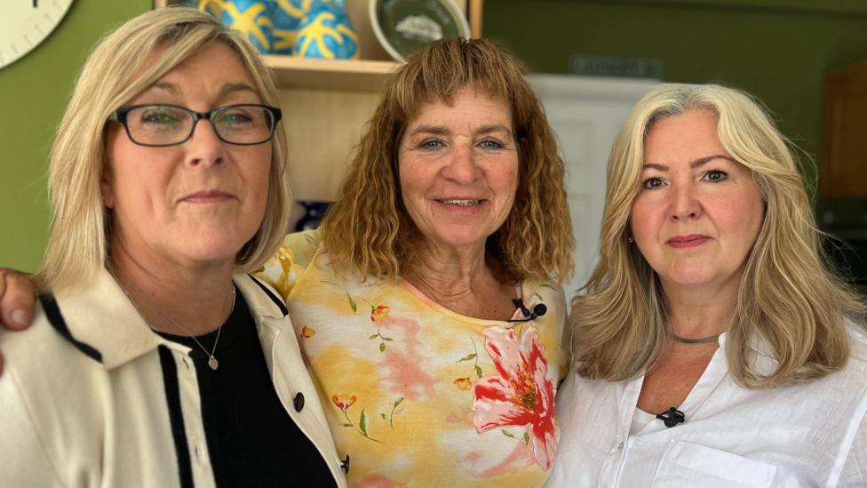 Left to right: Helen, Amanda and Joanne. Helen has blonde bobbed hair and is wearing glasses, a cream and white jacket and a silver pendant around her neck. Amanda has curly brown shoulder-length hair with a fringe and is wearing a yellow and pink floral top. Joanne has shoulder length blonde hair and is wearing a white shirt.