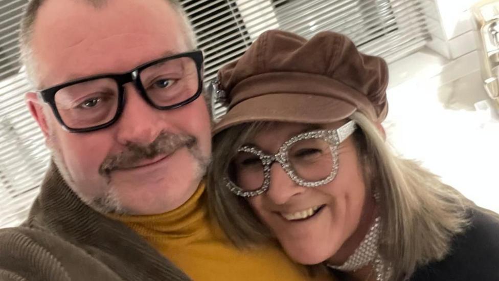 David Griffiths and Helen Moorefield at a fancy dress party. Helen is laughing and wearing large, sparkly diamante glasses. Griffiths is wearing a mustard turtle-neck jumper, brown, brown corduroy jacket and dark-rimmed glasses.