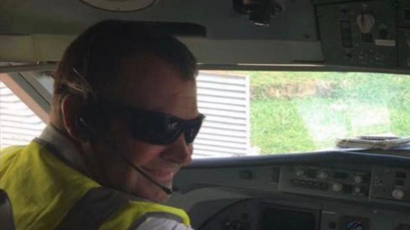Griffiths sitting in a helicopter wearing a fluorescent jacket, dark glasses and is wearing a headset microphone.