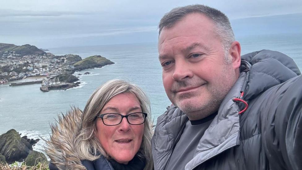 David Griffiths and Helen Moorefield dressed in outdoor clothing and standing by the sea. Both are smiling.