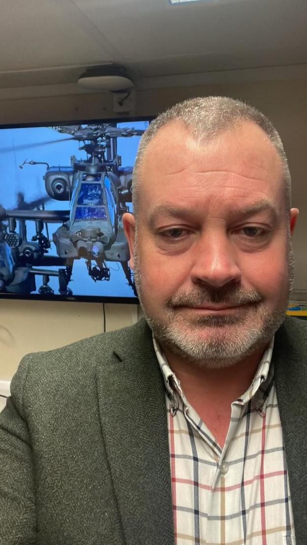 Griffiths dressed in a checked short and blazer standing in front of TV screen with a helicopter on it