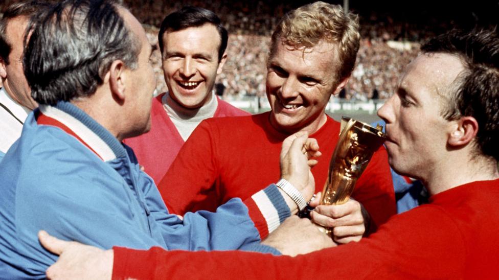 Sir Alf Ramsey in a light blue tracksuit (far left), Jimmy Armfield (centre left) with black hair in light turtle neck shirt and red sweater, Bobby Moore (centre right) with a red England shirt has blonde hair and Nobby Stiles (far right) has balck hair and red England shirt.