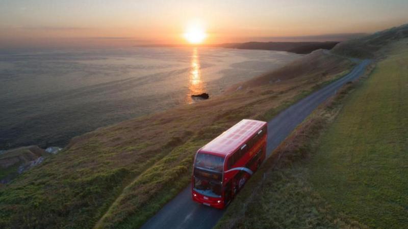 Red bus being driven along a quiet coastal road with the sun setting behind it.