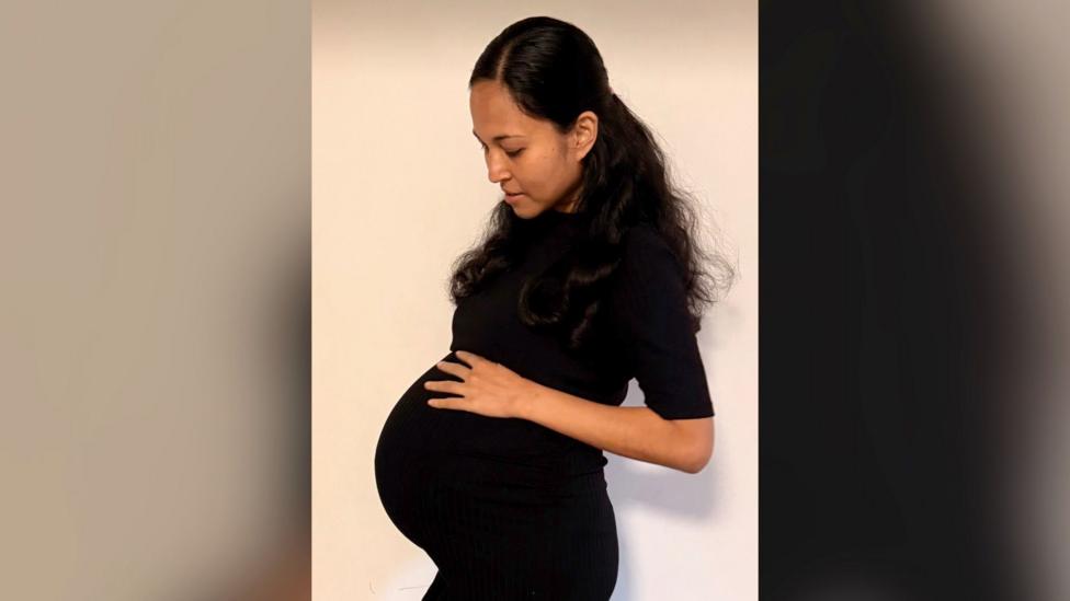 Jenitar Na'amoana pictured while pregnant. She has long dark hair and is wearing a black short-sleeved dress. She is looking down and touching a very visible baby bump.