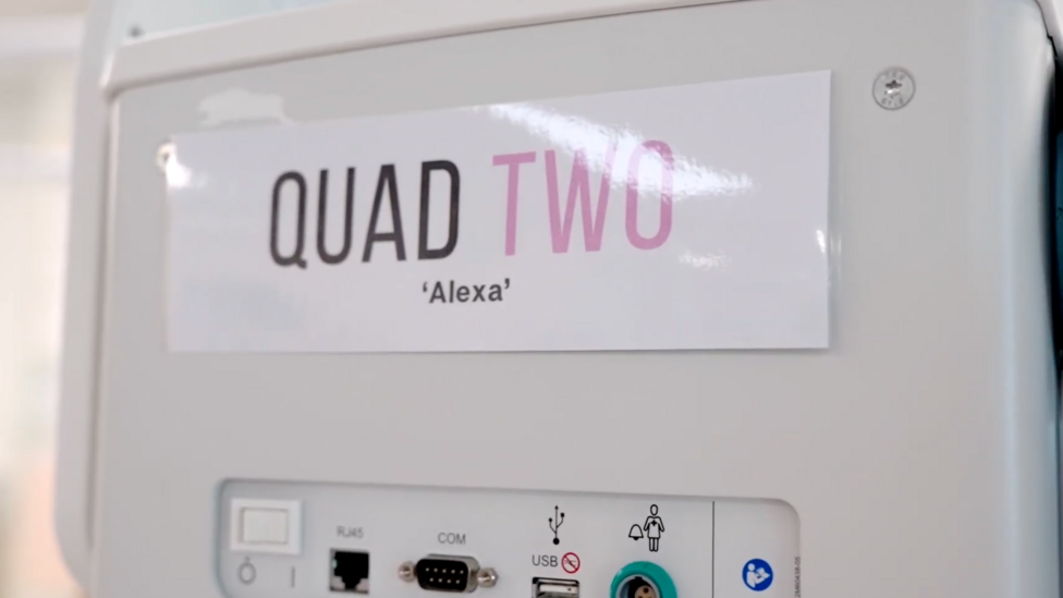 The side of a baby incubator with a laminated sign attached to it that reads 'Quad Two 'Alexa''