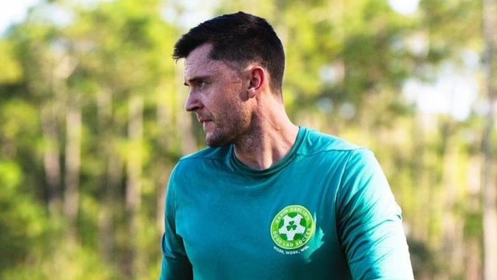 Coach Gavin Carlin has short brown hair and is wearing a green shirt and is looking off to the side.