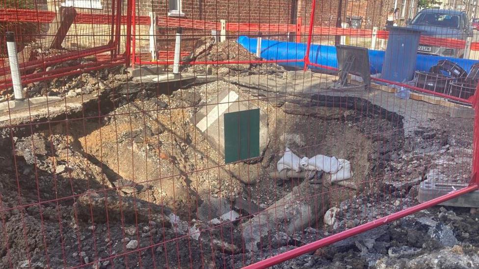 A hole in the ground. It is a couple of metres deep and filled with sand. There is red fencing around it.