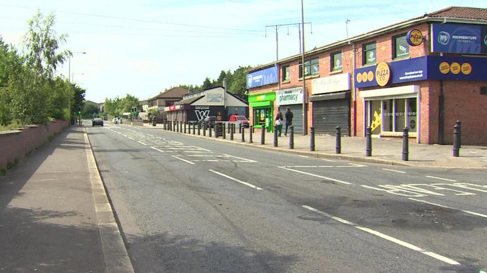 The Stewartstown Road, to the right are shops including a pizza shop and the left a wall and trees.