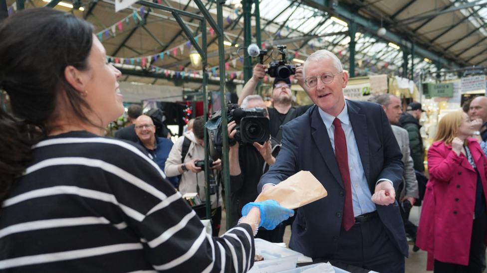 Hilary Benn who has grey hair and glasses and is wearing a suit is given a paper bag containing food by a woman stall holder with black hair at St George's Market