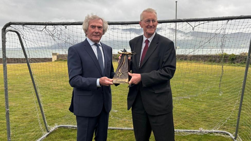 Pat Jennings has bushy grey hair and is wearing a suit and is jointly holding a trophy with Hilary Benn in front of a football net
