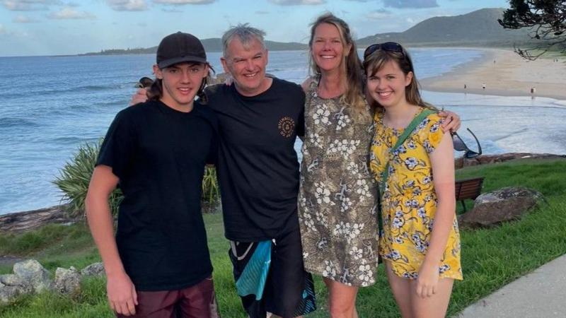 A family of four smile at the camera with a beach in the background