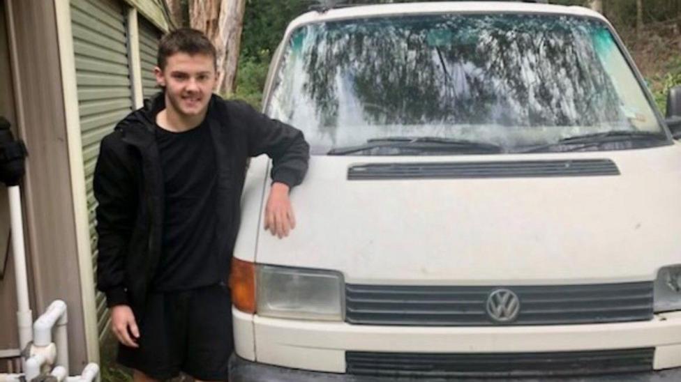 A teenage boy dressed in black stands next to a white van with his arm on the bonnet