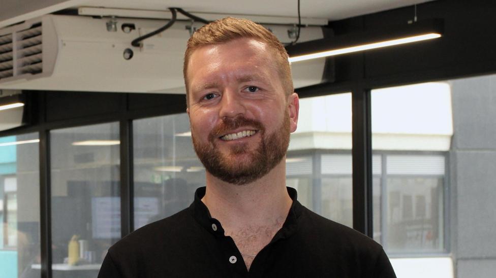 A smiling Chris Iveson wearing a black polo shirt looks straight into the camera.