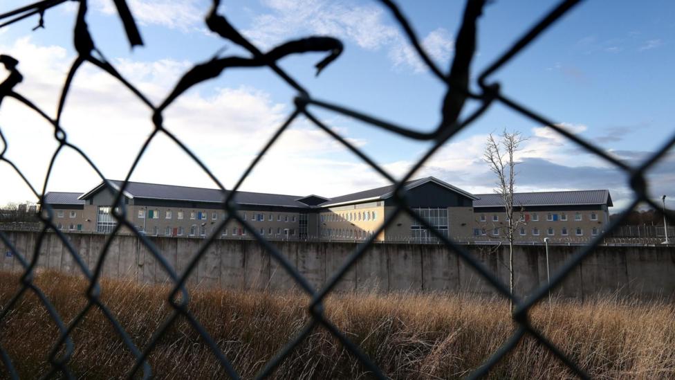 Low Moss Prison buildings behind a high wall, pictured through the wire of fence.