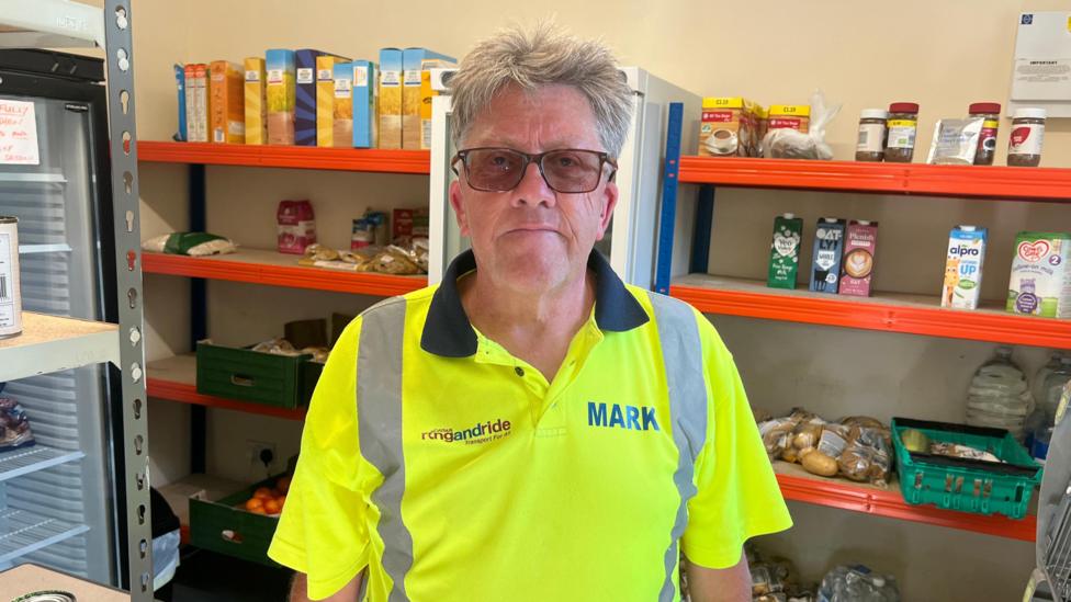 A man is wearing a high visibility polo shirt, with shelves of various food products behind him.